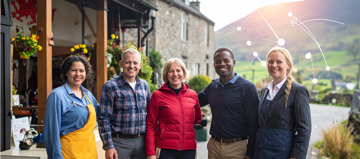 A diverse group of small business owners and entrepreneurs gathered outside a traditional Welsh shop, smiling and collaborating.